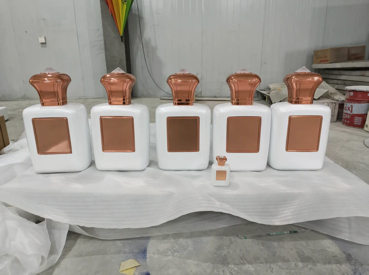 Five white 0.4 m factice perfume bottles with crackle lacquer bodies lined up on a bench beside the client's original bottle, all with blank copper plaques